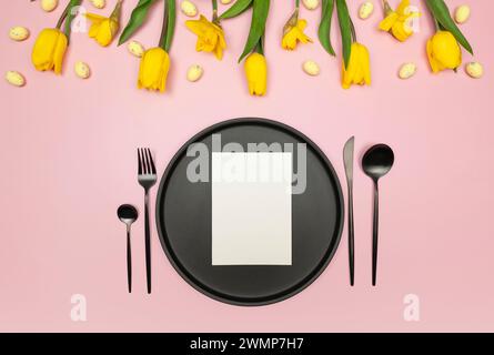 Vista dall'alto di tulipani gialli, narcisi, uova di pasqua su sfondo rosa. Tavolo, piatto nero e posate, carta bianca. Composizione primaverile di pasqua. Foto Stock