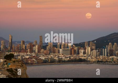 Lo skyline di Benidorm con Peñon de Ifach sullo sfondo e la luna piena al tramonto. Foto Stock