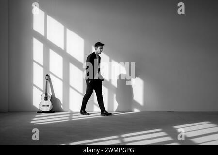 Un ragazzo in piedi si allontana dalla chitarra con le mani in tasca. Sta indossando un vestito nero. Interno di una ex fabbrica ristrutturata. Foto Stock