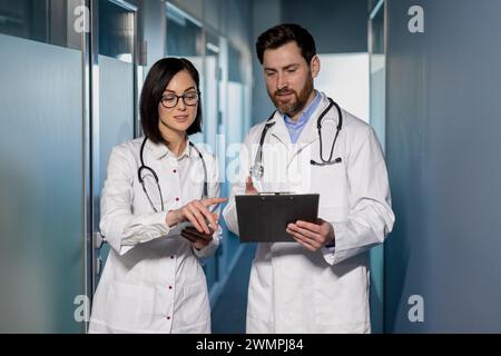 Due medici professionisti in camici da laboratorio bianchi che guardano gli appunti in mano mentre si trovano in sala clinica. Colleghi che discutono storia medica del paziente e formano una nuova strategia di trattamento. Foto Stock
