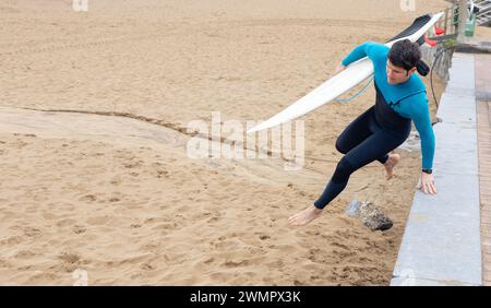 Un atletico surfista maschile in muta porta la sua tavola da surf bianca lungo la spiaggia, preparandosi ad entrare in mare per il surf. Foto Stock