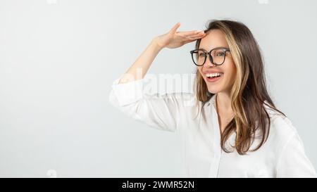 Una giovane donna esuberante con gli occhiali, che indossa una camicetta bianca, guarda in lontananza con la mano Foto Stock