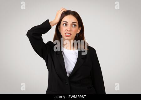 Una giovane donna d'affari sconcertata con un abito nero e una camicia bianca che si gratta la testa con un'espressione perplessa Foto Stock