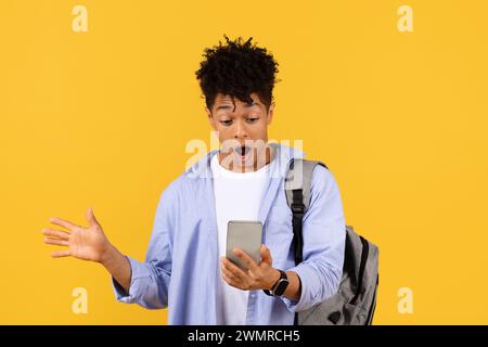 Uno studente maschio nero sorpreso che guarda uno smartphone con zaino su sfondo giallo Foto Stock