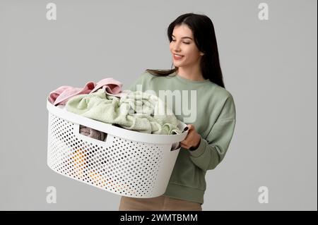 Bella giovane donna che tiene in mano un cesto con un bucato pulito su sfondo grigio Foto Stock