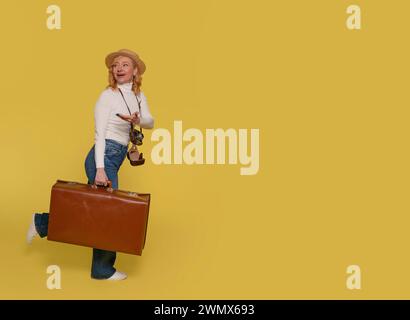 una donna in jeans, una camicia bianca e un cappello che porta una valigia e scatta foto con una macchina fotografica vintage su uno sfondo giallo. Gente felice che va avanti holida Foto Stock