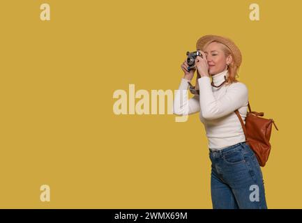 una donna in jeans, una camicia bianca e un cappello che porta una valigia e scatta foto con una macchina fotografica vintage su uno sfondo giallo. Gente felice che va avanti holida Foto Stock