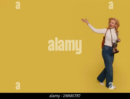 una donna in jeans, una camicia bianca e un cappello che porta una valigia e scatta foto con una macchina fotografica vintage su uno sfondo giallo. Gente felice che va avanti holida Foto Stock