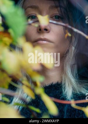 Primo piano del volto di una giovane donna bionda di platino caucasica pallida in contemplazione dietro un sottile fogliame autunnale colorato Foto Stock