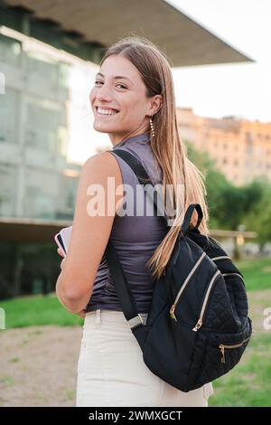 Vista posteriore verticale di una giovane studentessa felice che guarda la fotocamera godendosi i denti bianchi perfetti. Ritratto di una ragazza bionda gioiosa che posa per una promozione universitaria con un taccuino. Foto di alta qualità Foto Stock