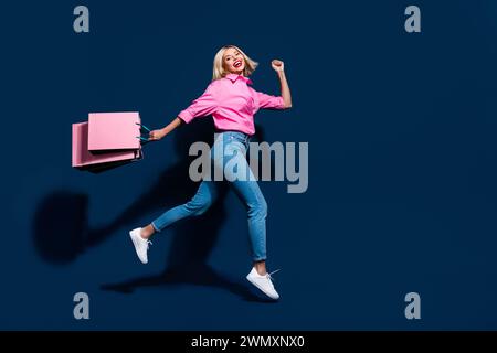 Foto a tutto il corpo di una giovane donna attraente che corre a portata di mano le borse della spesa indossano abiti rosa alla moda isolati su sfondo blu scuro Foto Stock