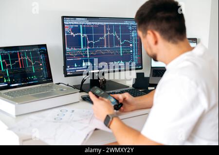 Il trader maschile siede in ufficio davanti a schermi con grafici e grafici finanziari, utilizzando smartphone, predice, analizza e studia la crescita o il declino della criptovaluta.Borsa, trading Foto Stock