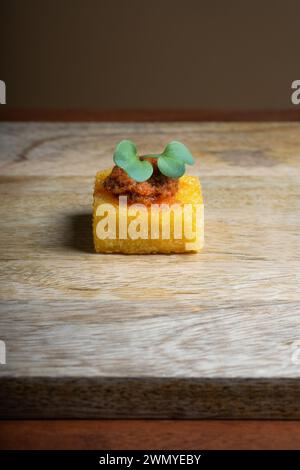 Una canape di polenta gourmet condita con un ricco stufato di pomodoro e cipolla e un rametto di fresca decorazione verde, elegantemente presentato su un asse di legno. Foto Stock