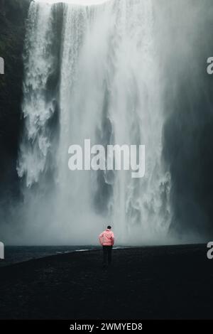 La vista posteriore di un uomo irriconoscibile con una giacca rosa si erge osservando la maestosa cascata Skogafoss in Islanda, circondata da nebbia e scura vulcanica Foto Stock