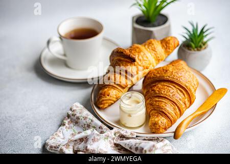 Una colazione con due croissant dorati su un piatto, accompagnati da un barattolo di miele e una tazza di tè, con piccole piante in vaso nel dorso Foto Stock