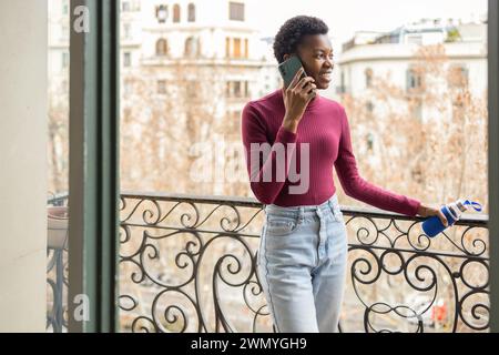 Giovane donna nera in un abbigliamento casual che parla su uno smartphone mentre si trova su un balcone con sfondo urbano. Foto Stock