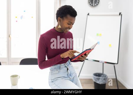 Una allegra donna afroamericana è impegnata con un tablet digitale, in un ambiente di ufficio luminoso con una lavagna chiara e un orologio nella b Foto Stock