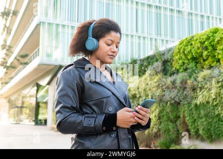 Una giovane donna con un cappotto nero usa il suo smartphone indossando le cuffie blu, in piedi di fronte a un edificio moderno con cespugli verdi Foto Stock