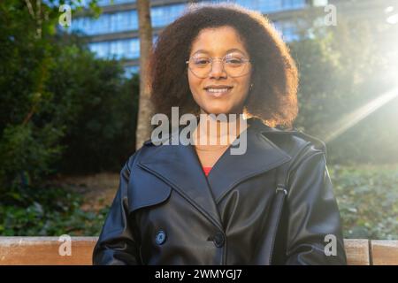 Giovane donna radiosa in occhiali sorridente su una panchina del parco con la luce del sole che filtra tra tra gli alberi e un edificio sullo sfondo Foto Stock