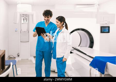 Due membri del personale medico, un uomo e una donna, sono impegnati in una discussione su un tablet digitale in una sala radiologica ben attrezzata con uno scanner MRI Foto Stock