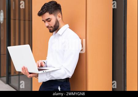 Un giovane uomo d'affari concentrato con una camicia bianca brillante lavora intensamente sul suo notebook contro una parete moderna dai toni caldi Foto Stock