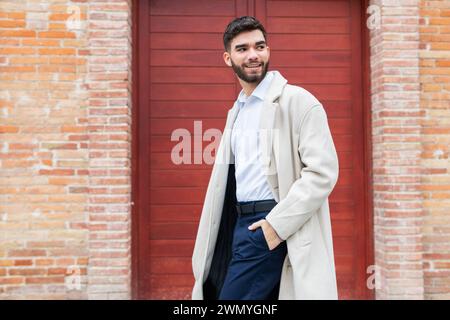 Un giovane uomo elegante in abbigliamento da lavoro sorride con fiducia mentre si trova sullo sfondo di una porta rossa, trasudando un senso di professionalità informale nel suo Foto Stock