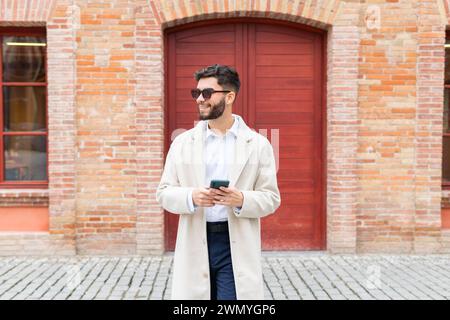 Allegro uomo d'affari con un trench e occhiali da sole che navigano sul suo telefono, davanti a un classico edificio in mattoni con una porta rossa Foto Stock
