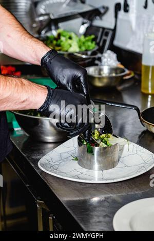 Primo piano delle mani di uno chef con guanti neri che ripiano delicatamente un'insalata gourmet in un anello di metallo su un piatto con motivi di marmo in una cucina professionale Foto Stock
