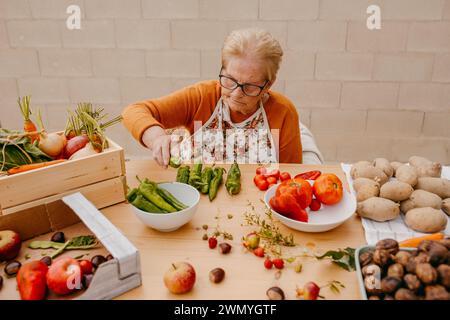 Una donna anziana con i bicchieri trita meticolosamente le verdure in una cucina ben illuminata, circondata da una vasta gamma di prodotti sani Foto Stock