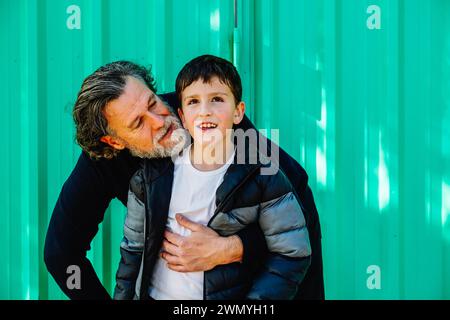 Una foto emozionante che cattura un bambino sorridente abbracciato dal suo affettuoso padre su uno sfondo verde vivace Foto Stock