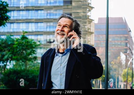Un uomo d'affari maturo ama conversare sul suo cellulare, con edifici di uffici sullo sfondo, mostrando la fusione tra tecnologia e uso quotidiano Foto Stock