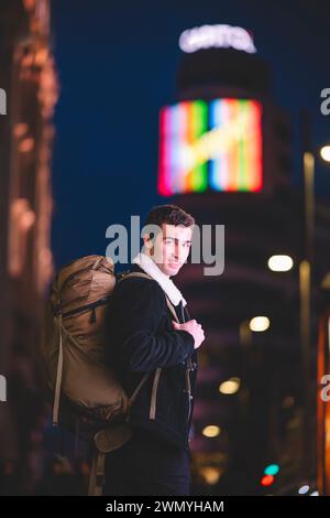 Un giovane sorridente con uno zaino in una strada illuminata al neon di Madrid, che guarda la macchina fotografica Foto Stock