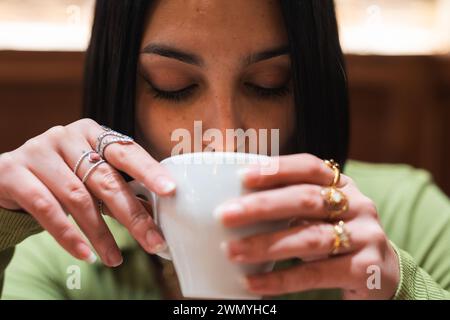 Una giovane donna dai capelli scuri e decorata con anelli delicati sorseggia una tazza da caffè bianca, un momento intimo catturato in un ambiente interno accogliente. Foto Stock