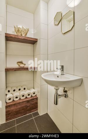 Un piccolo bagno contemporaneo con piastrelle in ceramica bianca, scaffalature in legno, un lavandino in porcellana bianca e specchi esagonali sulle pareti. Foto Stock