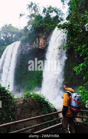 Vista laterale del viaggiatore da solo con telefono cellulare che scatta foto mentre ammiri le maestose cascate di Iguazu da un sentiero lussureggiante nel nord dell'Argentina. Foto Stock