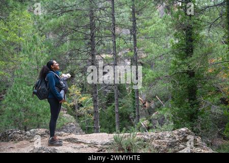 Una donna felice con un bambino in un portabagagli si erge tra alti pini, godendosi un momento tranquillo in un'escursione nella natura Foto Stock