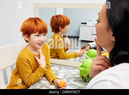 Una logopedista che lavora e allena un bambino dai capelli rossi con difficoltà di parola per sviluppare le capacità di linguaggio, linguaggio e comunicazione Foto Stock