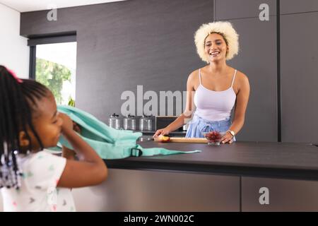 La giovane madre birazziale sorride mentre prepara il pranzo scolastico, la figlia afroamericana osserva Foto Stock