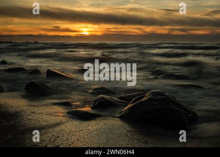 Nel tranquillo abbraccio di Veczemju Klintis, il sole che tramonta dipinge una scena affascinante sulla spiaggia rocciosa, catturata con eterea bellezza per lungo tempo Foto Stock