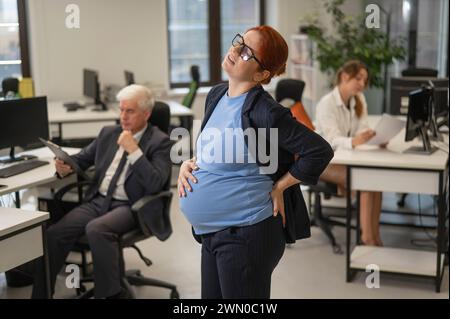 Una donna incinta soffre di dolore e tiene lo stomaco in piedi nel mezzo dell'ufficio accanto ai suoi colleghi. Foto Stock
