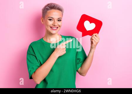 Ritratto di una ragazza allegra sorriso di toothy indica che il braccio del dito è tenuto come un'icona di notifica, uno spazio vuoto isolato su sfondo rosa Foto Stock