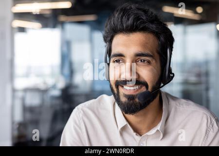 Foto ravvicinata del volto di un giovane indiano che indossa una cuffia che lavora online mentre è seduto in ufficio, sorride e guarda di lato. Foto Stock