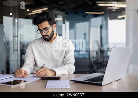 Un giovane indiano attento e serio lavora come contabile, revisore dei conti e analista finanziario. Sedersi alla scrivania in ufficio e lavorare con account e dati. Foto Stock