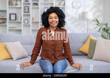 Ritratto di una giovane donna afroamericana vestita con abiti casual seduta sul divano a casa, sorridente e guardando la macchina fotografica. Foto Stock