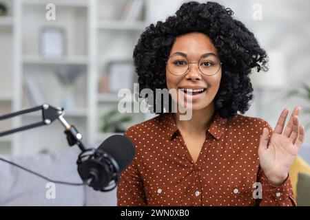 Una giovane donna afroamericana sta registrando una trasmissione online, insegnando da remoto, conversando in uno studio domestico e andando dal vivo. Si siede sul divano di fronte alla telecamera e saluta con un sorriso. Foto Stock