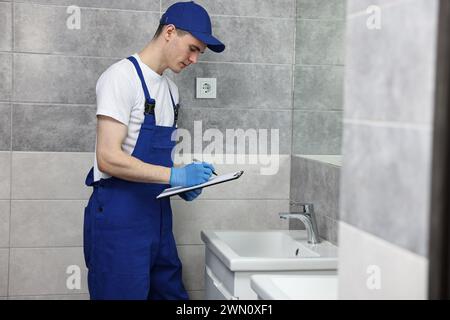 Giovane idraulico che scrive i risultati dell'esame del rubinetto in bagno Foto Stock