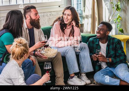 Un gruppo di amici allegro e multiculturale condivide risate con bicchieri di vino rosso e una ciotola di popcorn in un accogliente soggiorno illuminato dal sole Foto Stock