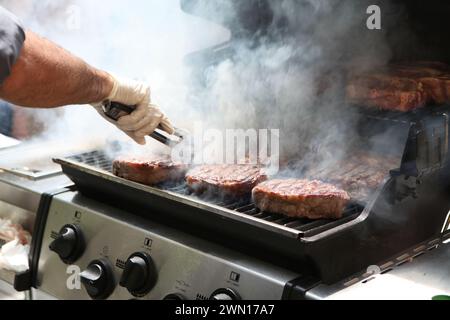 Primo piano della mano di un uomo che tiene un paio di pinze, trasformando bistecche di manzo su un barbecue affumicato Foto Stock