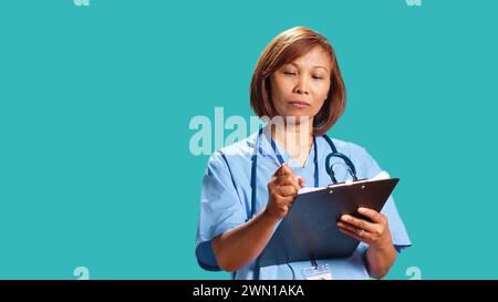 Primo piano di un esperto di assistenza sanitaria che scrive i dati degli appuntamenti dei pazienti in arrivo sugli appunti mentre sono al lavoro. Infermiere BIPOC isolato su sfondo blu studio finendo i documenti Foto Stock