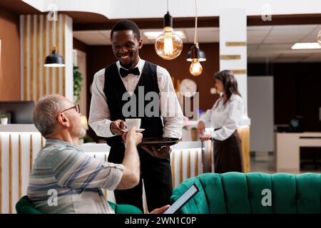 Dipendente afroamericano che serve caffè a turisti anziani con tablet alla reception dell'hotel di lusso. Senior man che tiene in mano un dispositivo digitale e riceve una tazza di tè dal cameriere del resort nell'area lounge. Foto Stock